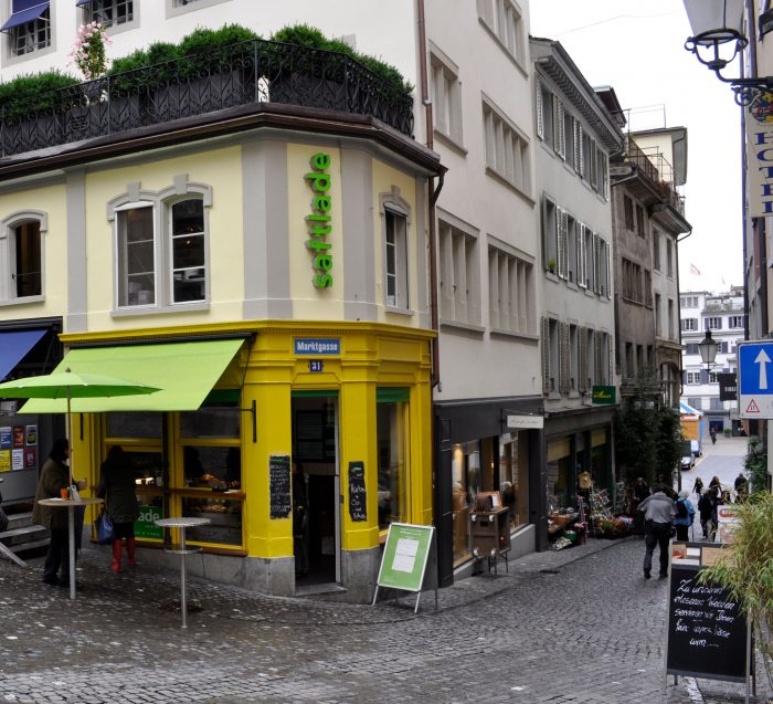 Gelbes Eckhaus mit Saftladen und Stehtischen in einer Altstadtgasse mit Kopfsteinpflaster