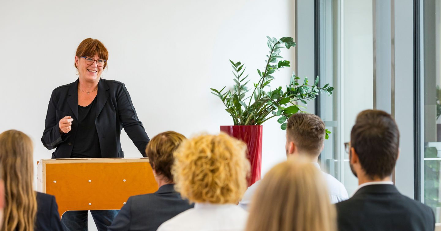 Referentin hält am Rednerpult einen Vortrag vor Publikum in einem hellen Seminarraum