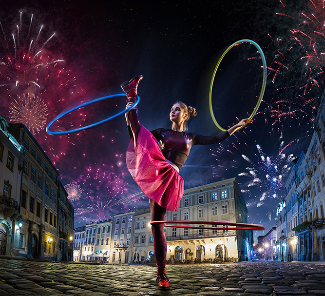 Artistin mit Hula-Hoop-Reifen auf einem nächtlichen Altstadtplatz vor Feuerwerk