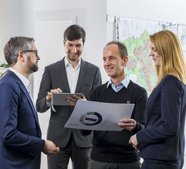 Vier Beratende besprechen ein Diagramm und ein Tablet vor einer Wandkarte