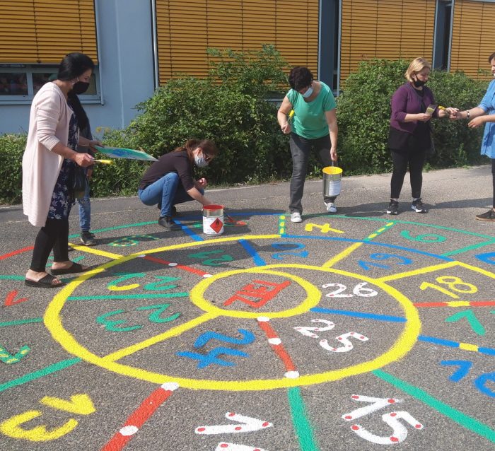 Frauen mit Mund-Nasen-Schutz malen ein buntes Zahlenspielfeld auf den Asphalt eines Hofs