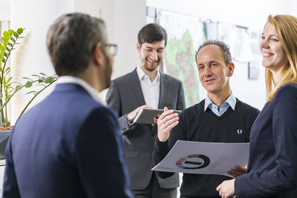 Vier Mitarbeitende im Gespräch im Büro, einer hält ein Blatt mit Kreisdiagramm